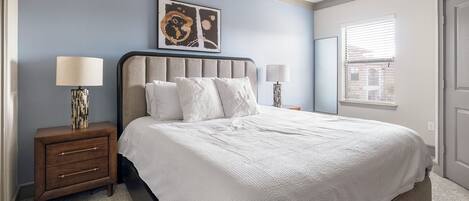 A bedroom with a large bed, two bedside tables, a wall-mounted artwork, and a window with blinds.