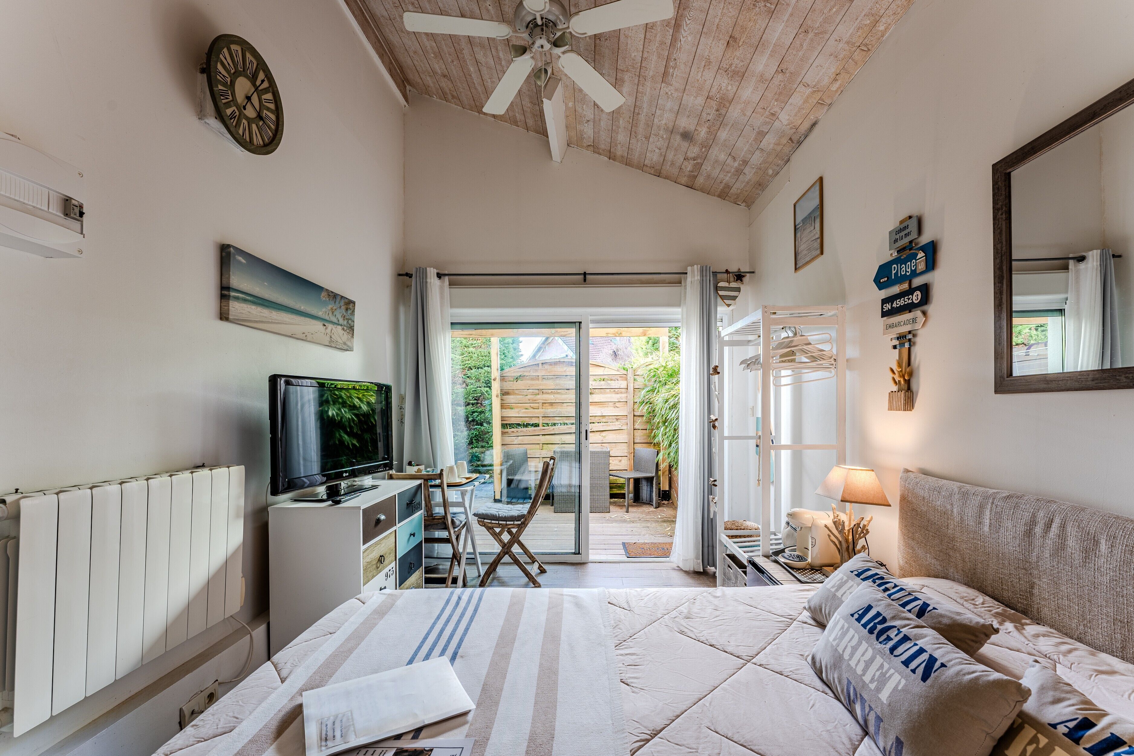 A bedroom with a bed, a television, a ceiling fan, a radiator, a desk, and a view of the outside.