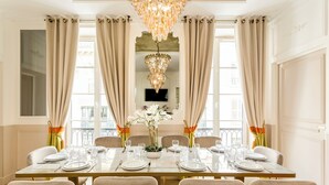 Una sala da pranzo formale con un lungo tavolo apparecchiato per sei persone, ampie finestre con tende, un lampadario e un televisore a schermo piatto montato a parete.