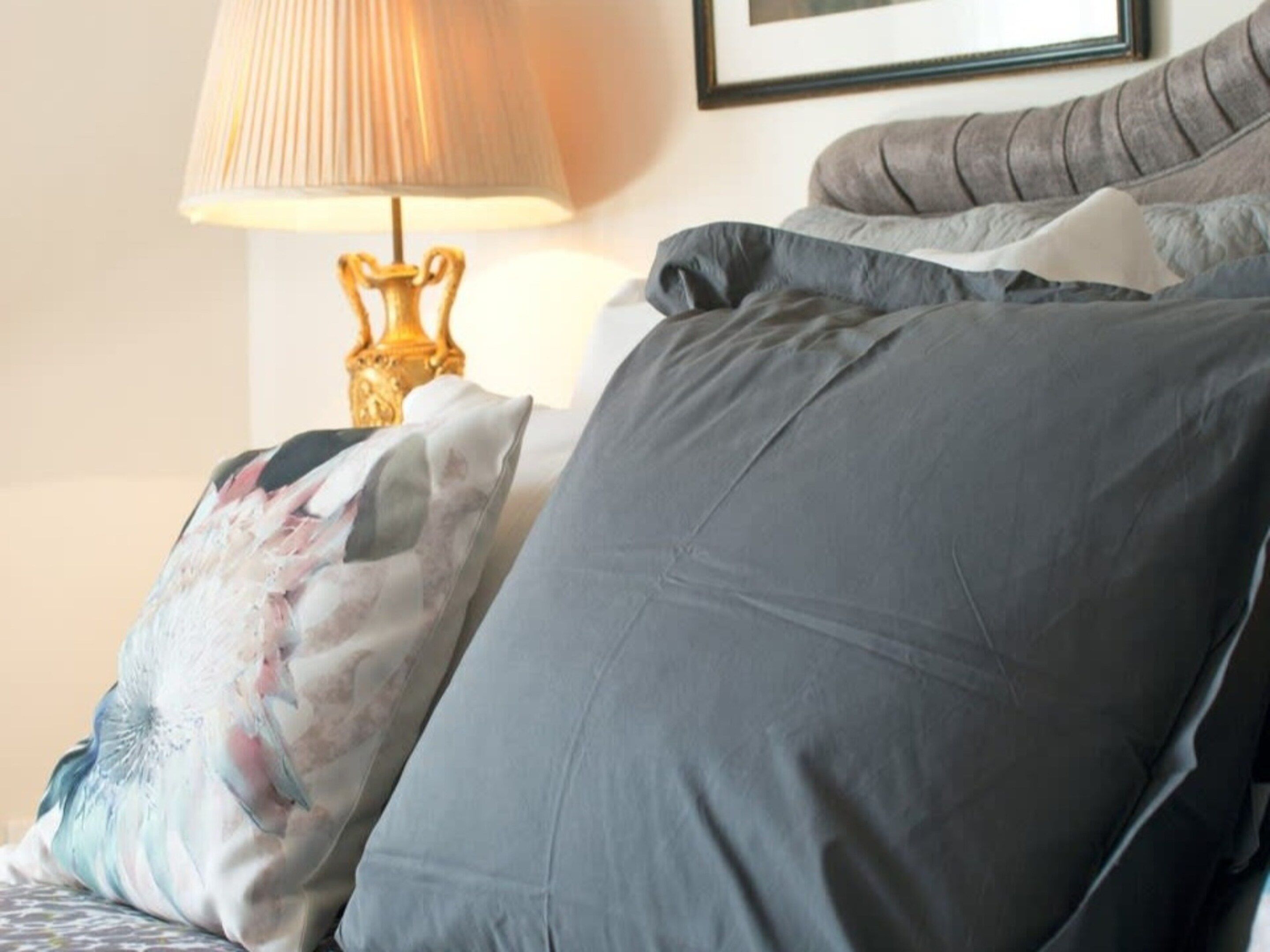 A bed with grey and patterned pillows, a lamp with a golden base, and a framed picture on the wall.