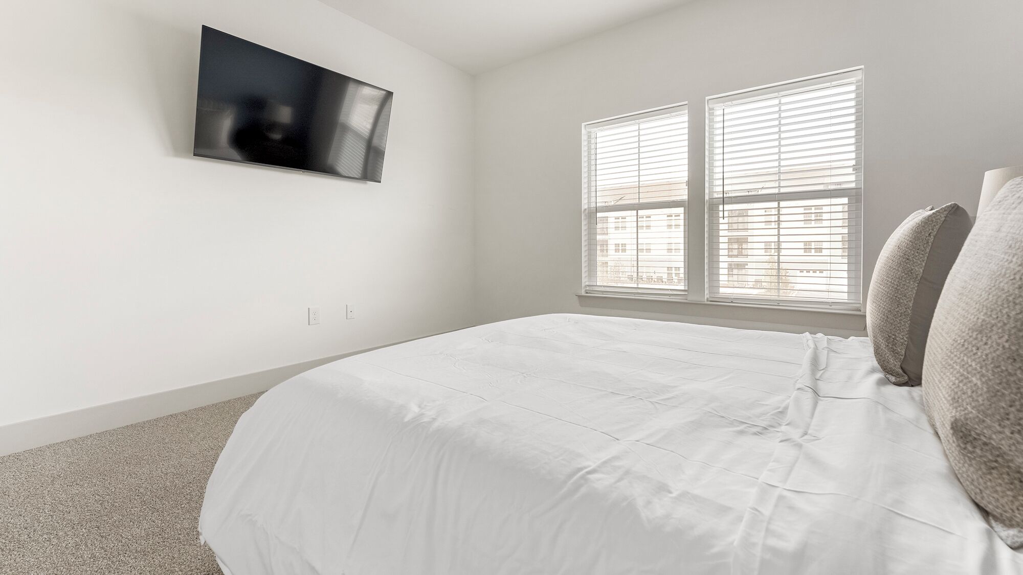 A bedroom with a bed, a TV mounted on the wall, and a window with blinds.