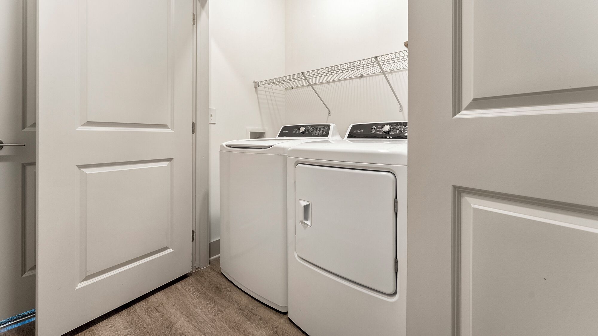 Una sala de lavandería con dos lavadoras y secadoras apiladas frontalmente, un tendedero metálico sobre las secadoras, y una puerta que da a otra habitación.
