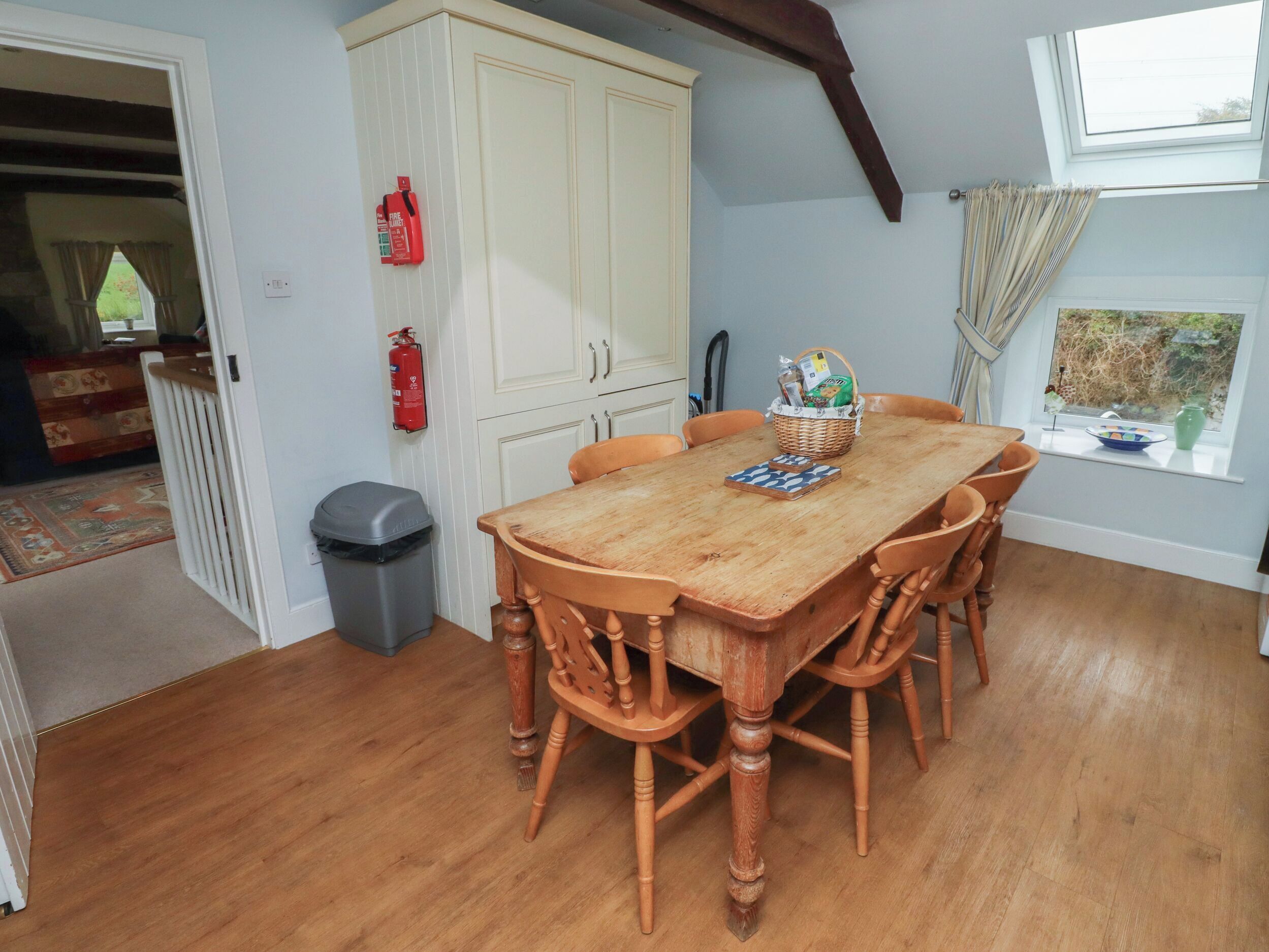 Un espace repas comprenant une table et des chaises en bois, un extincteur, une poubelle et une fenêtre avec des rideaux.