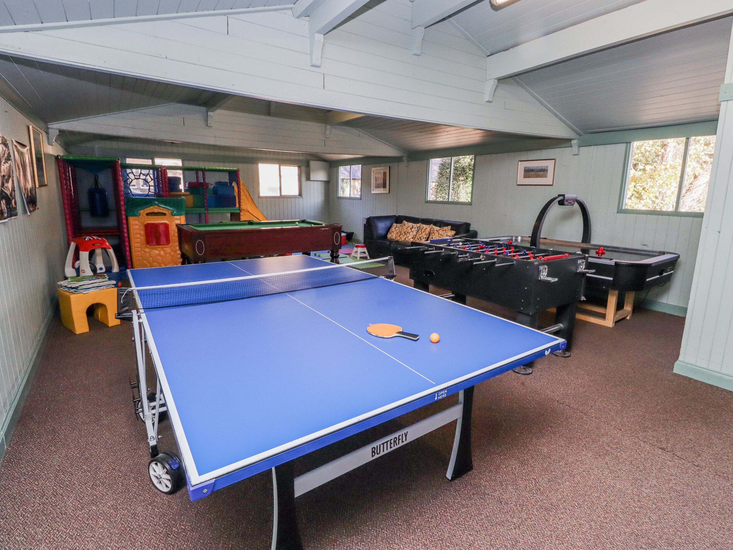 A room with a ping pong table, foosball table, and a pool table. There are windows and a ceiling with exposed beams.