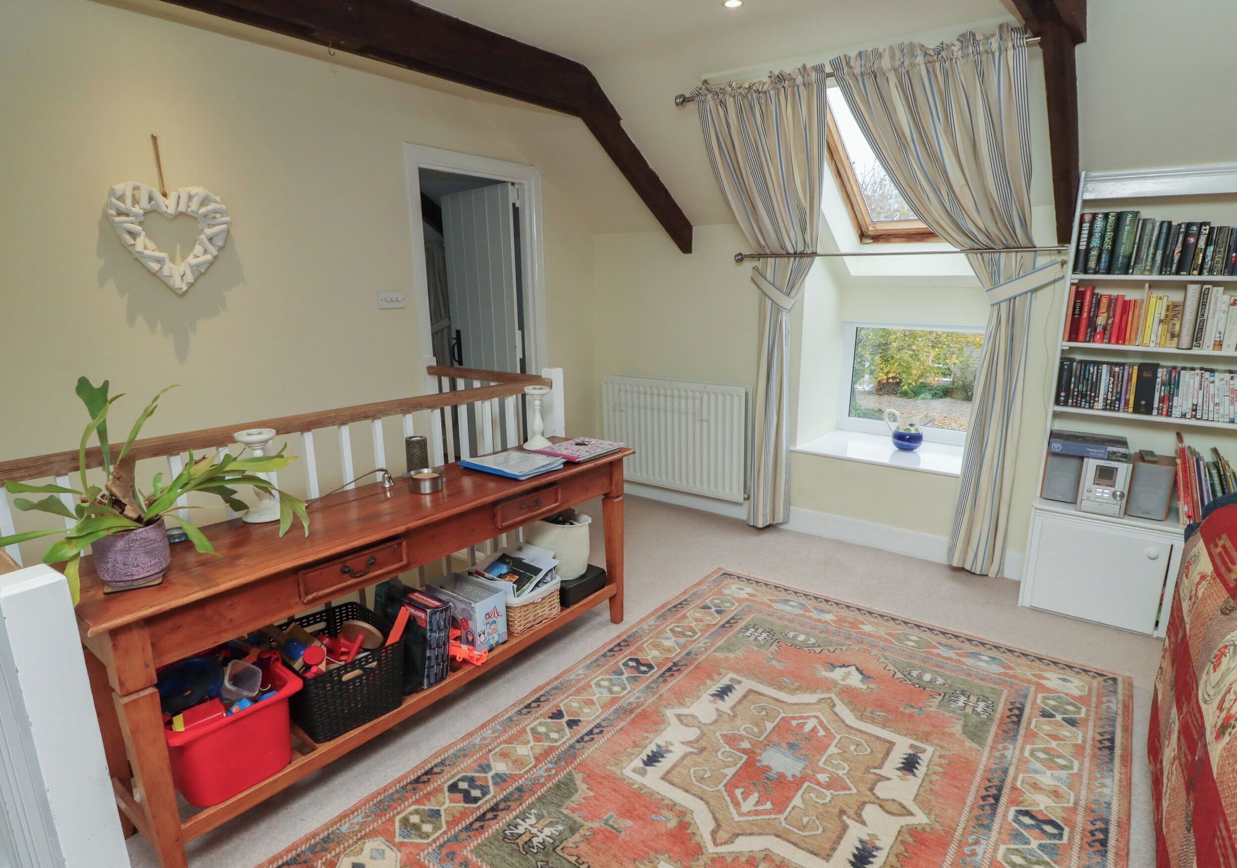 A cozy living room with a wooden bench, a bookshelf, a patterned rug, and a view through a window with curtains.