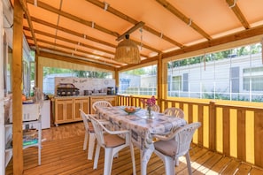 A covered outdoor dining area with a wooden deck, a table with chairs, a hanging light fixture, and a view of a mobile home in the background. - Mobile Home 'Location Agde' with Private Terrace, Wi-Fi and Air Conditioning (Agde)