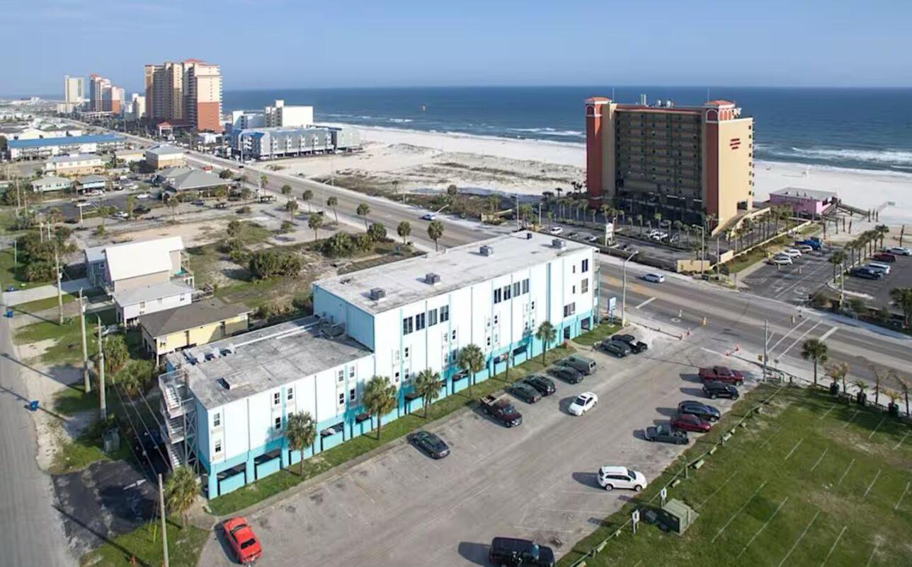 Vista aérea de una zona costera con un gran edificio hotelero, estacionamientos y una playa al fondo.