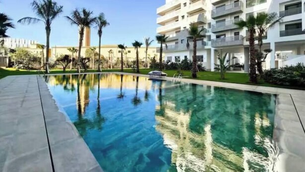 Un hotel moderno con piscina, rodeado de palmeras y un cielo azul despejado.