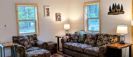 A cozy living room with a wooden ceiling, two sofas with patterned cushions, a red and beige rug, and a small table with a lamp.