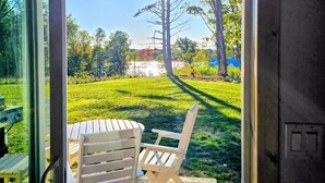 Eine Terrasse mit Tisch und zwei Stühlen, eine Glastür zu einem Rasen mit Bäumen und einem wolkenlosen Himmel.