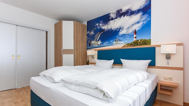 A neatly made bed with white linens, a blue headboard, and a painting of seagulls and a lighthouse on the wall.