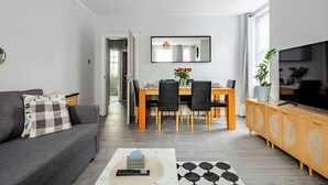 Una sala moderna con un sofá gris, una mesa de comedor con sillas, un televisor sobre un mueble de madera y un espejo grande en la pared.