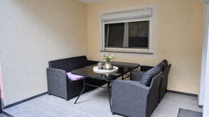 Een klein terras buiten met een tafel en vier stoelen, een raam erboven en een deur aan de rechterkant.