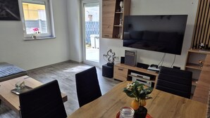 Een moderne woonkamer met een flatscreen televisie, houten meubels, een eettafel met stoelen en een potplant.