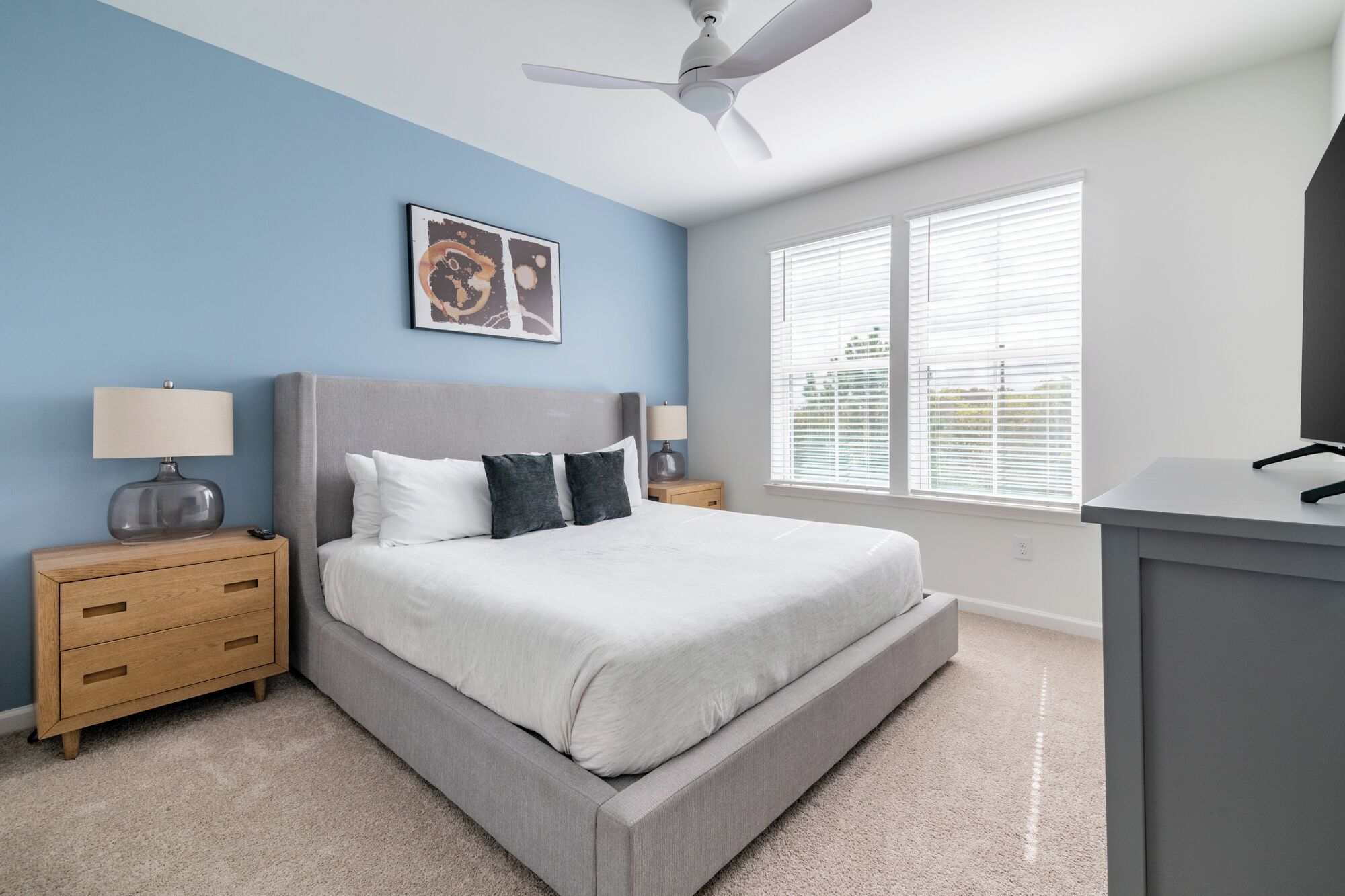 A bedroom with a bed, bedside tables, a ceiling fan, a window with blinds, and a wall-mounted TV.