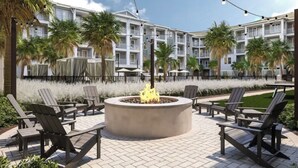 A courtyard with a circular fire pit surrounded by Adirondack chairs, palm trees, and a building with balconies in the background. - Landing Apartments Downtown Palm Coast (Palm Coast)