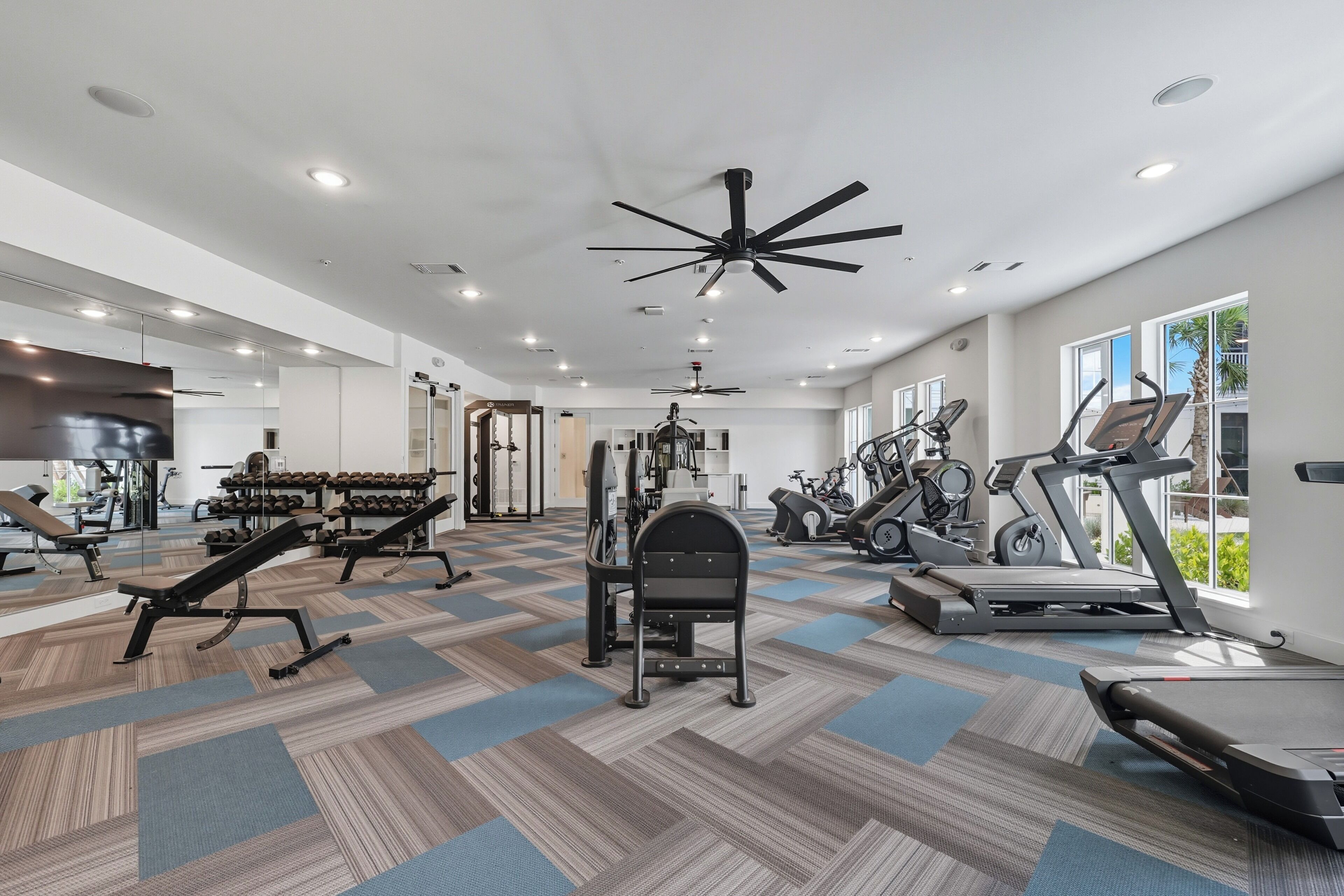 Une salle de sport moderne équipée de matériel d’exercice, de ventilateurs de plafond et de grandes fenêtres.