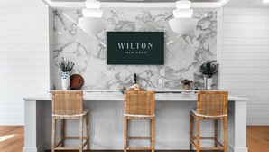 A modern kitchen with a marble backsplash, three wicker bar stools, and a framed sign that reads "Wilton Palm Coast". - Landing Apartments Downtown Palm Coast (Palm Coast)