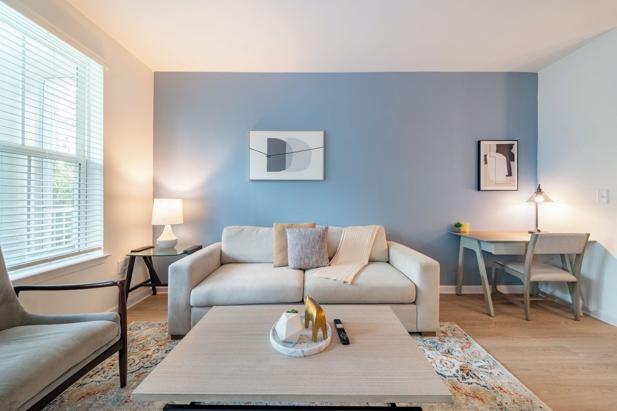 Un salon moderne avec un canapé de couleur claire, une table basse agrémentée d’objets décoratifs, une table d’appoint avec une lampe et un bureau avec une chaise.