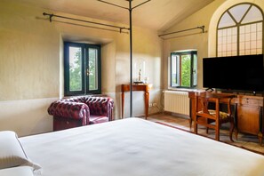 A hotel room with a large bed, a desk with a television, a red leather chair, and two windows with views of greenery. - 'Ficaiola' Room with Shared Pool and Garden (Massa Marittima)