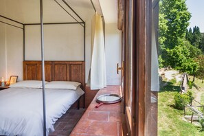 Une chambre à coucher avec un lit à baldaquin, une tête de lit en bois, une table de chevet avec une lampe, et une vue sur un jardin par une porte ouverte.