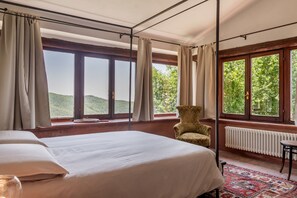 A room with a large bed, a chair, a radiator, and a view of greenery through large windows.