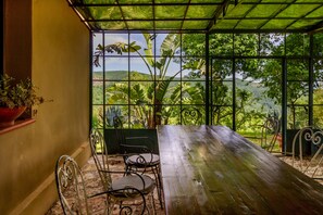 A wooden table and chairs under a green roof with a view of lush greenery and hills.