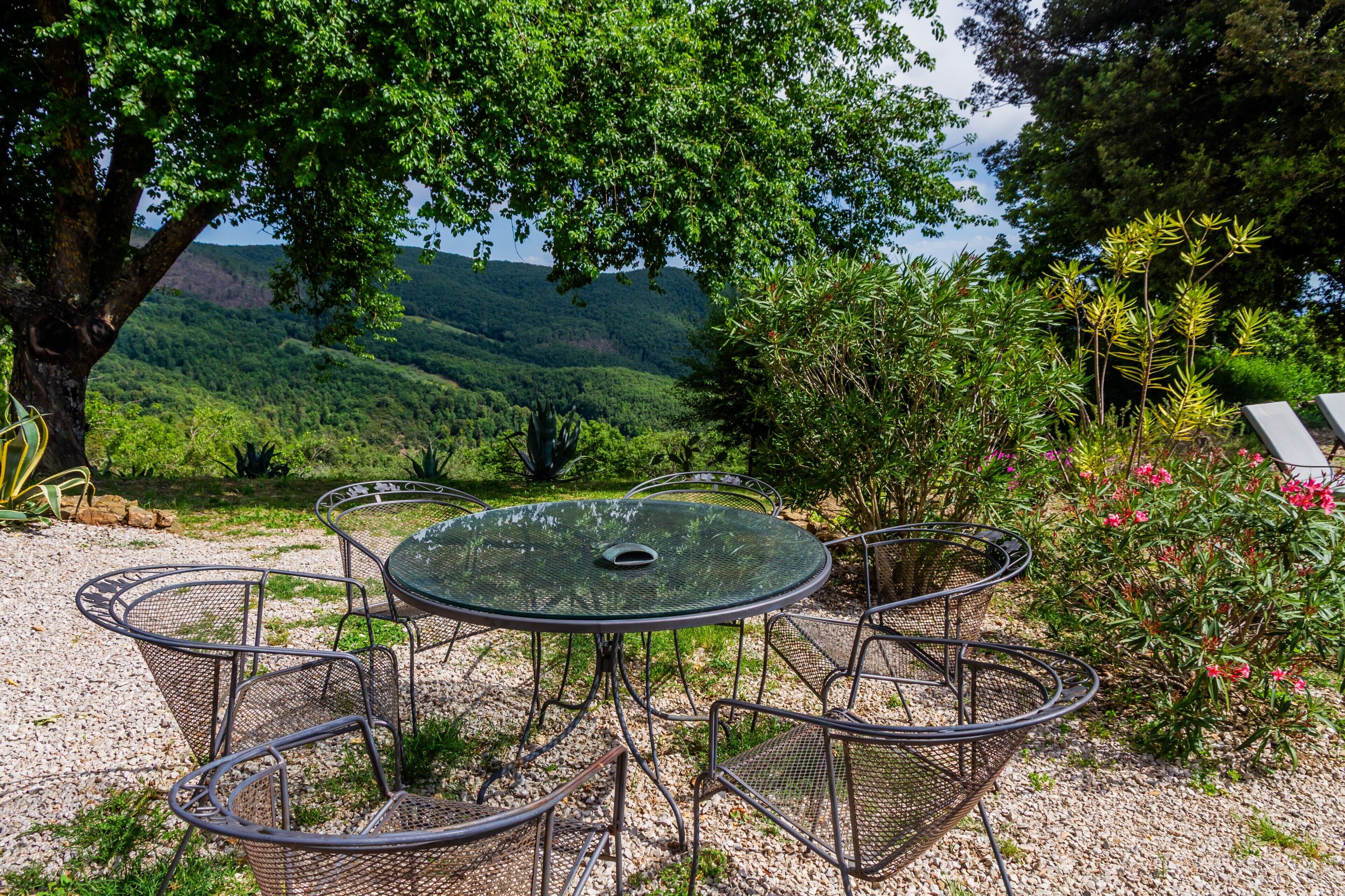 An outdoor seating area with a round table and chairs, surrounded by greenery and a view of a hilly landscape.