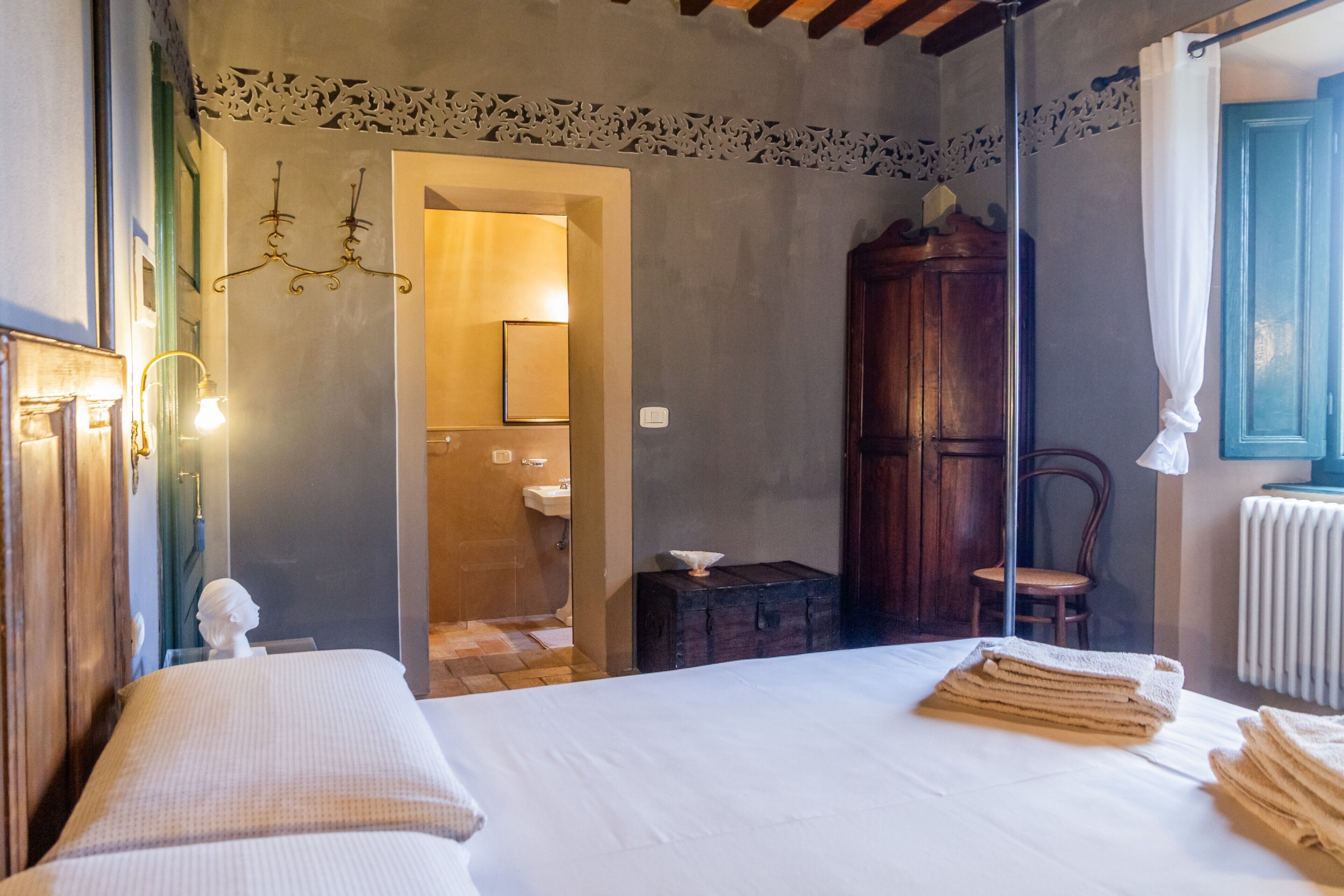 A bedroom with a bed, a wooden wardrobe, a chair, a small table, and a bathroom visible through an open door.