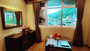A room with a desk, chair, and a table with a vase of flowers. A large window offers a view of a mountainous area. - Keswani Group Tashi Heritage Hotel (Gangtok)
