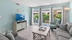 A living room with a TV, a wooden coffee table, a sofa, and a chair. There is a balcony with a view of greenery and a building. - Includes Beach Service! ⛱️Walk to Beach-Pool+Hot tub-Fitness Area-Sea Oats 211☀️ (Fort Walton Beach)