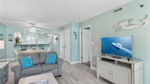 A living room with a grey sofa, a coffee table, a TV on a white cabinet, and a ceiling fan. - Includes Beach Service! ⛱️Walk to Beach-Pool+Hot tub-Fitness Area-Sea Oats 211☀️ (Fort Walton Beach)