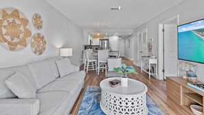 Ett modernt vardagsrum med en grå soffa, ett runt soffbord med en växt och en stor TV som visar en vy över kusten.