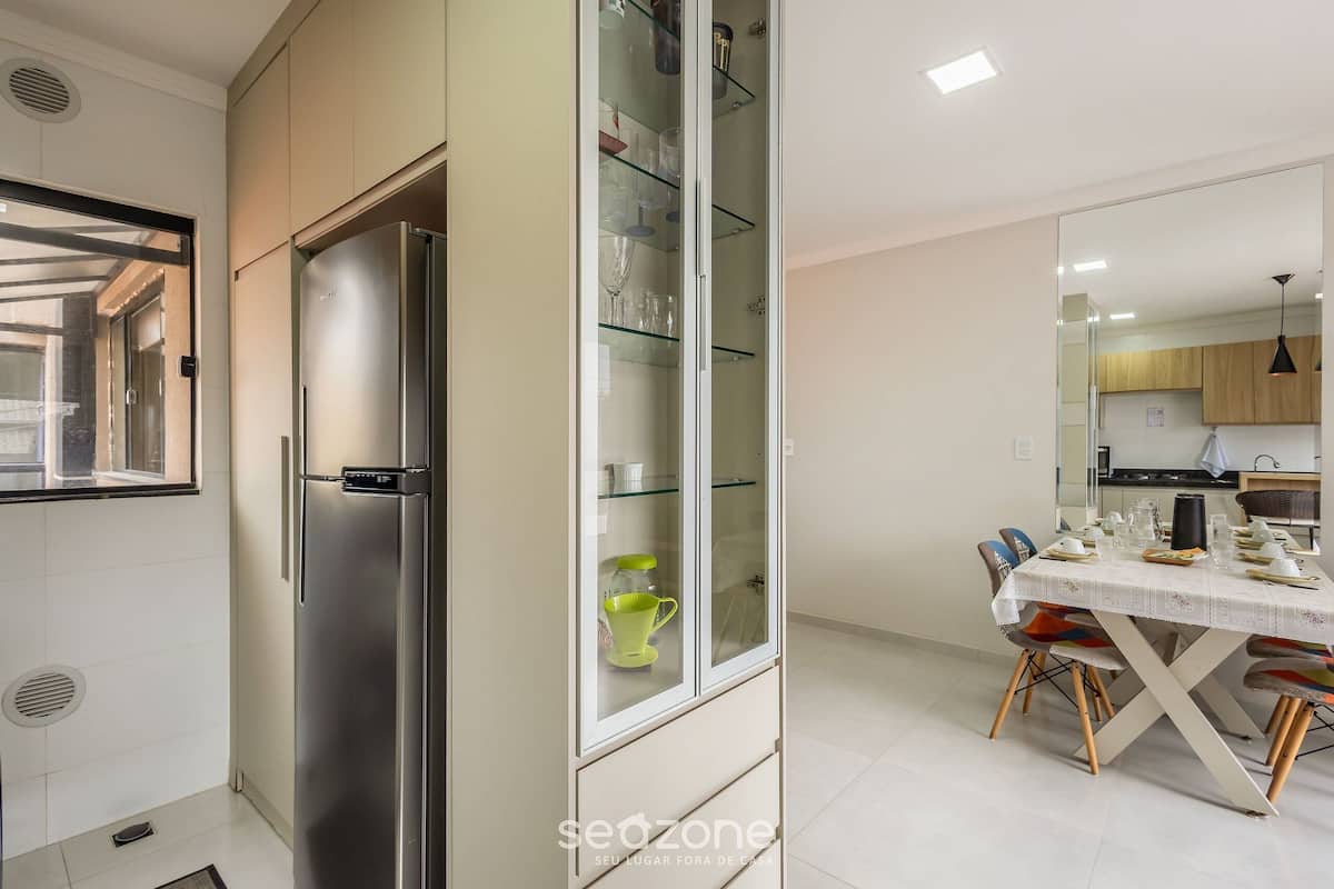 Una cocina moderna con un refrigerador de acero inoxidable, un armario con puerta de vidrio que contiene vajilla, y un comedor con una mesa lista para dos personas.