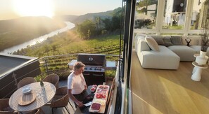 A balcony with a grill, dining table, and chairs, overlooking a river and hills.