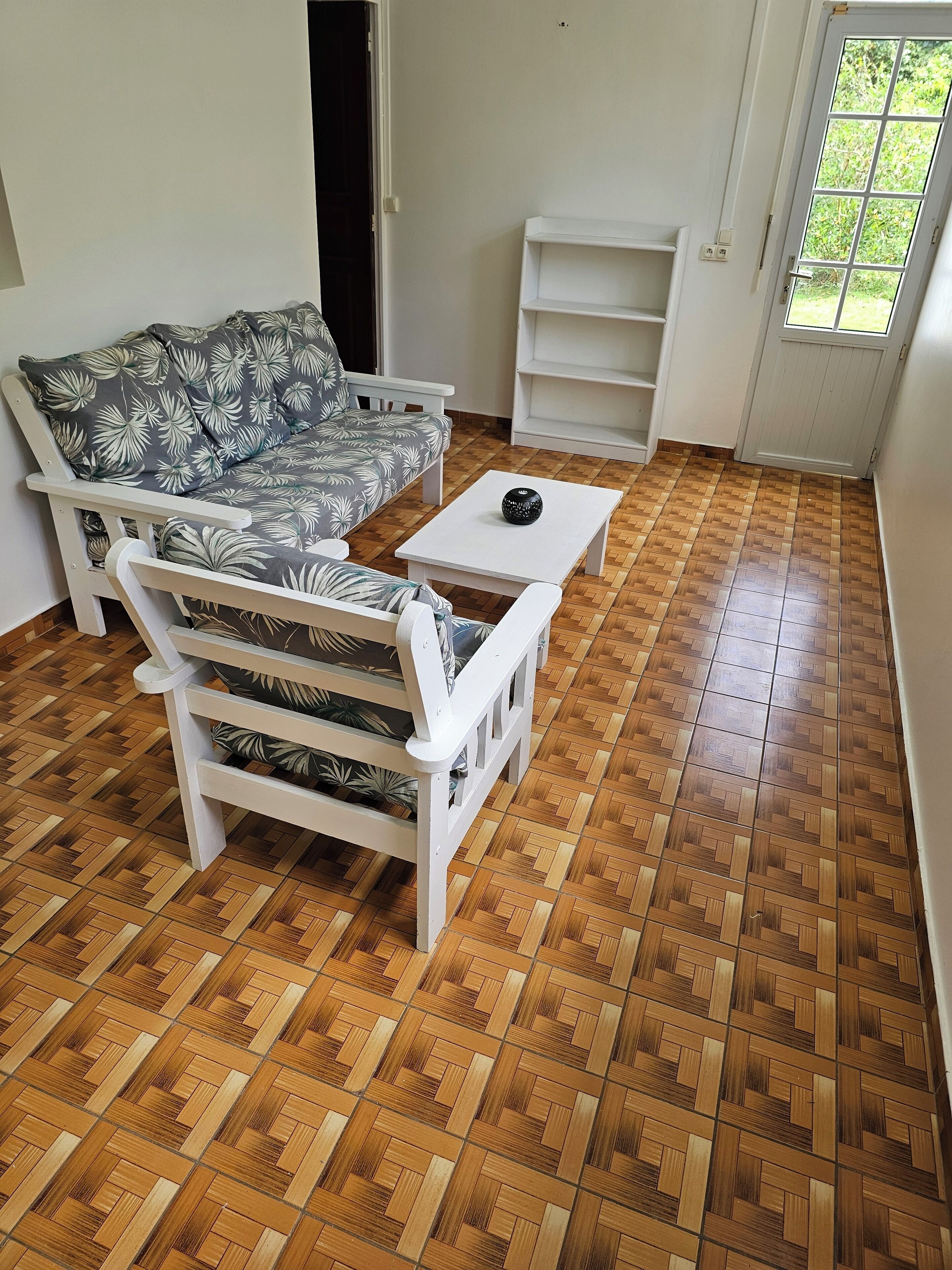 A room with a patterned sofa, a white armchair, a small white table, and a herringbone wooden floor.