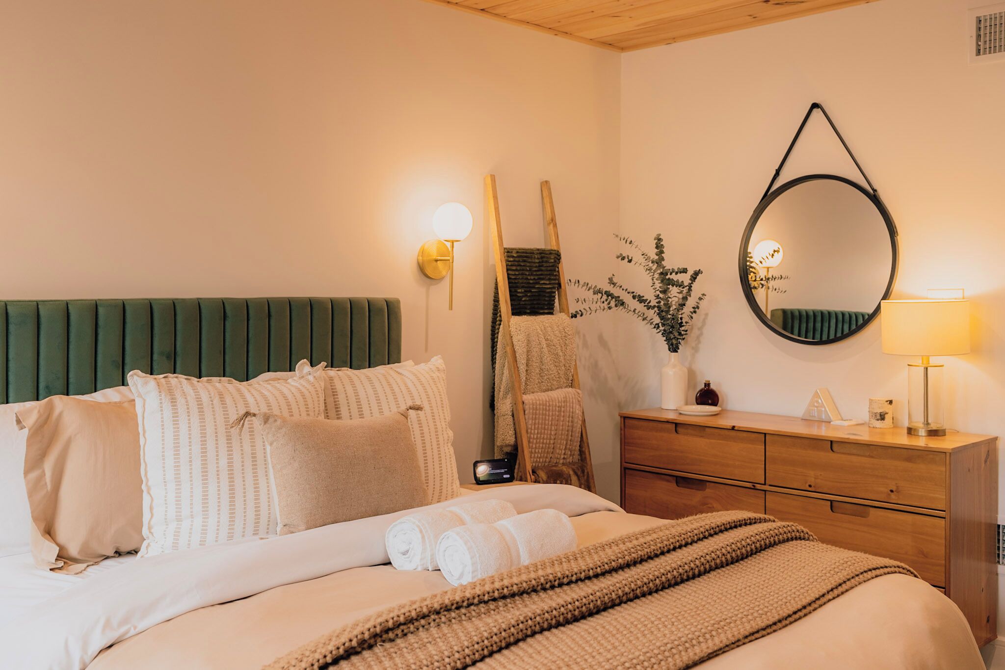 Uma cama bem arrumada com almofadas, uma luminária de cabeceira, um espelho e um aparador de madeira.