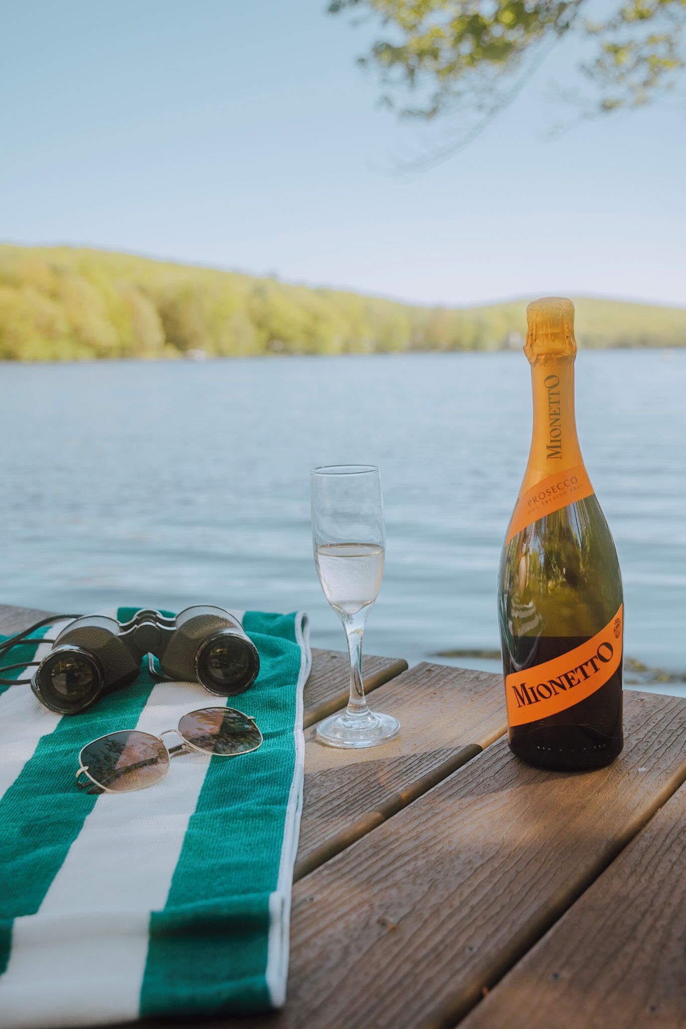 Una botella de Monetto Prosecco sobre una mesa de madera, junto a un vaso de agua y unas gafas de sol.
