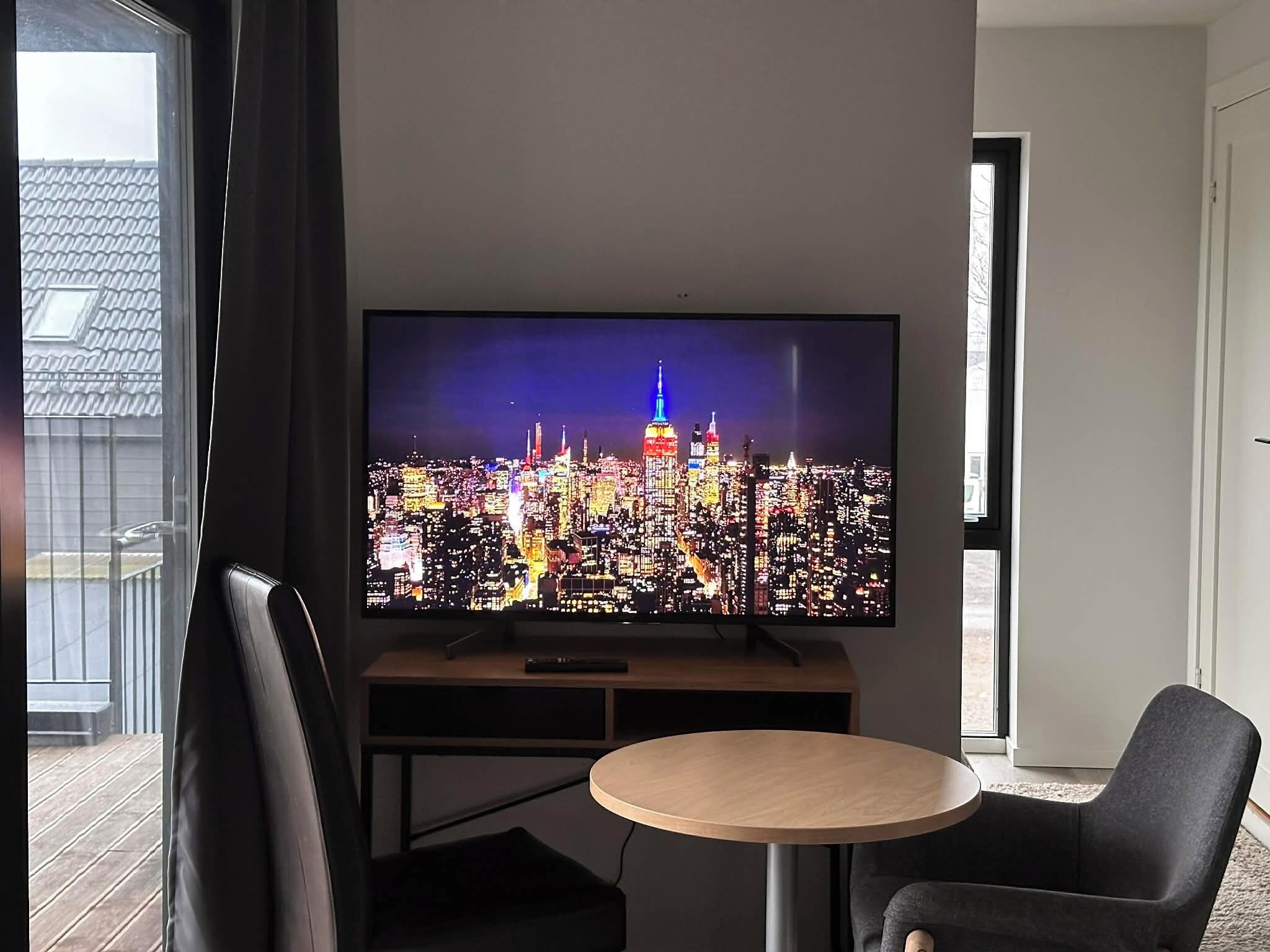 Una sala de estar moderna con una televisión de pantalla plana mostrando un paisaje urbano, una mesa redonda de madera y dos sillas negras.