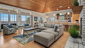 A spacious living room with a large sectional sofa, a coffee table, and patterned armchairs. A kitchen area with a white countertop and hanging plants is visible in the background. - This Cape Cod style beauty will make you fall in love with the Forgotten Coast! (Alligator Point)