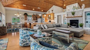 A spacious living room with a high ceiling, wooden beams, a fireplace, and a kitchen in the background. - This Cape Cod style beauty will make you fall in love with the Forgotten Coast! (Alligator Point)