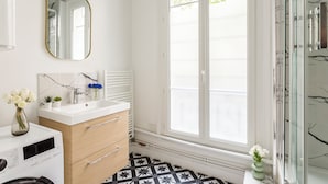 A modern bathroom with a white vanity, a mirror, a radiator, and a window with a view of greenery. - Family Suite - Marais Turbigo I (Paris)