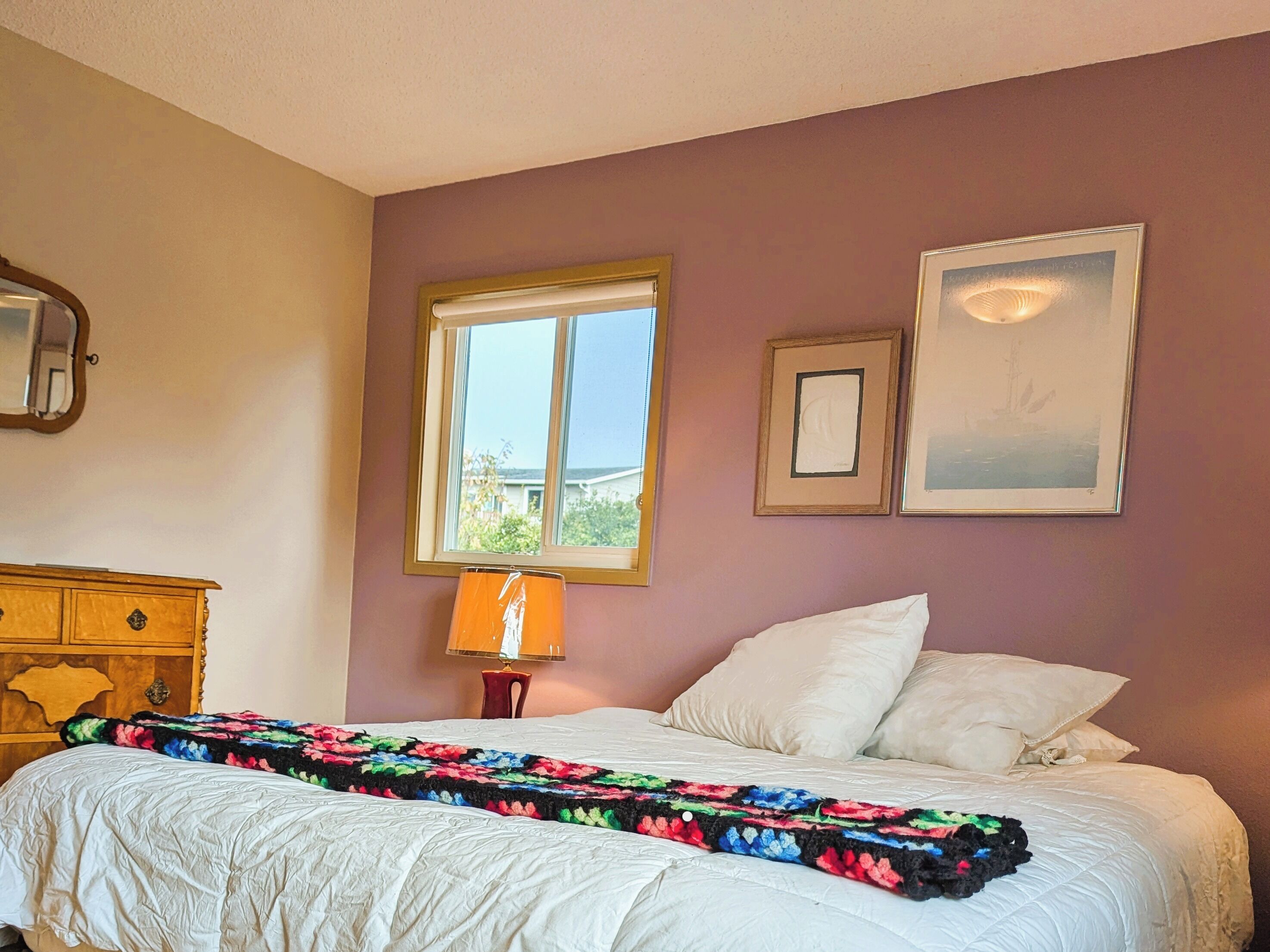A bedroom with a bed, a wooden dresser, a window, and two framed pictures on the wall.