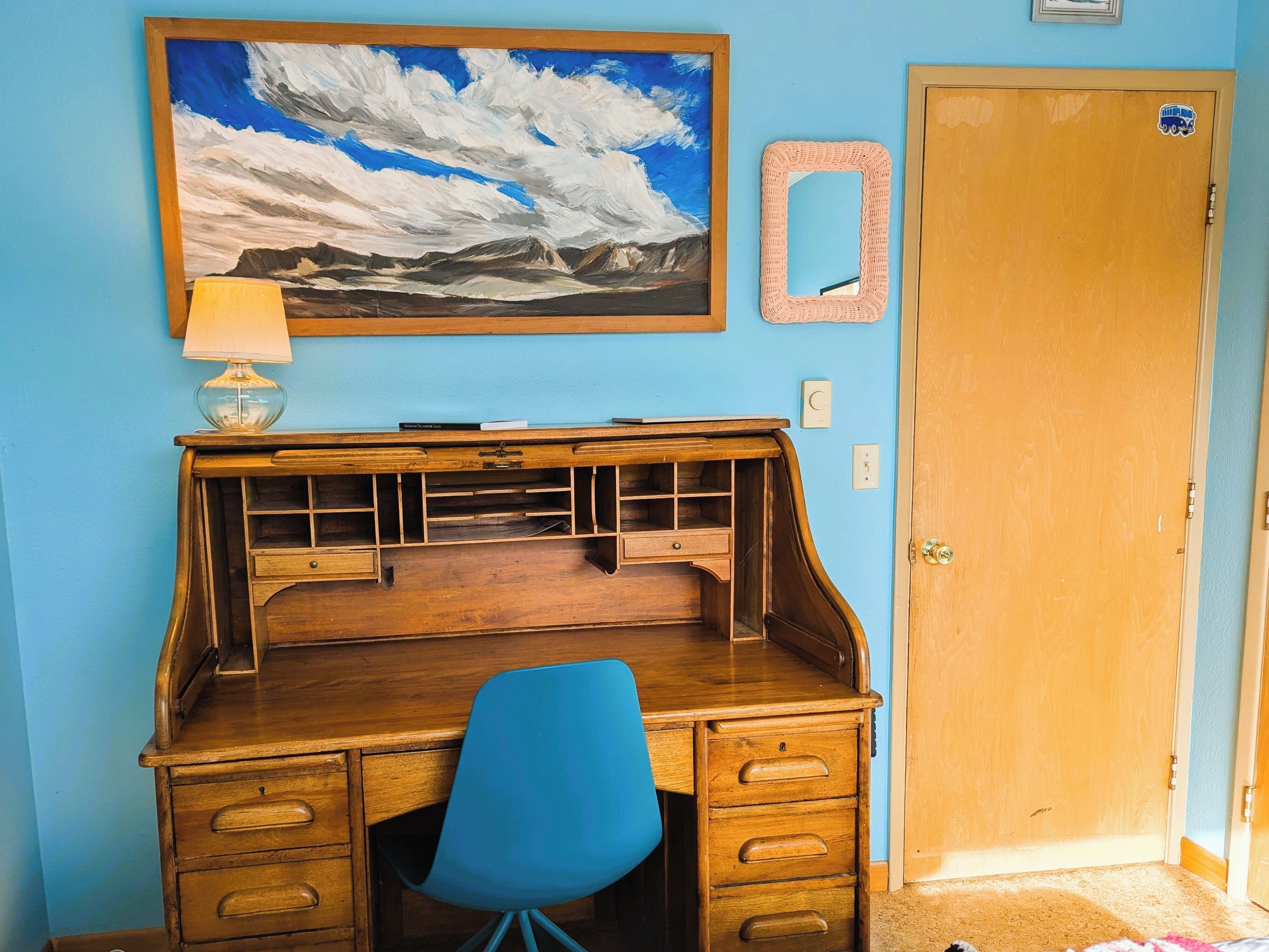 Un escritorio de madera con cajones, una silla azul, un cuadro enmarco con un paisaje de montaña, un espejo y una puerta.