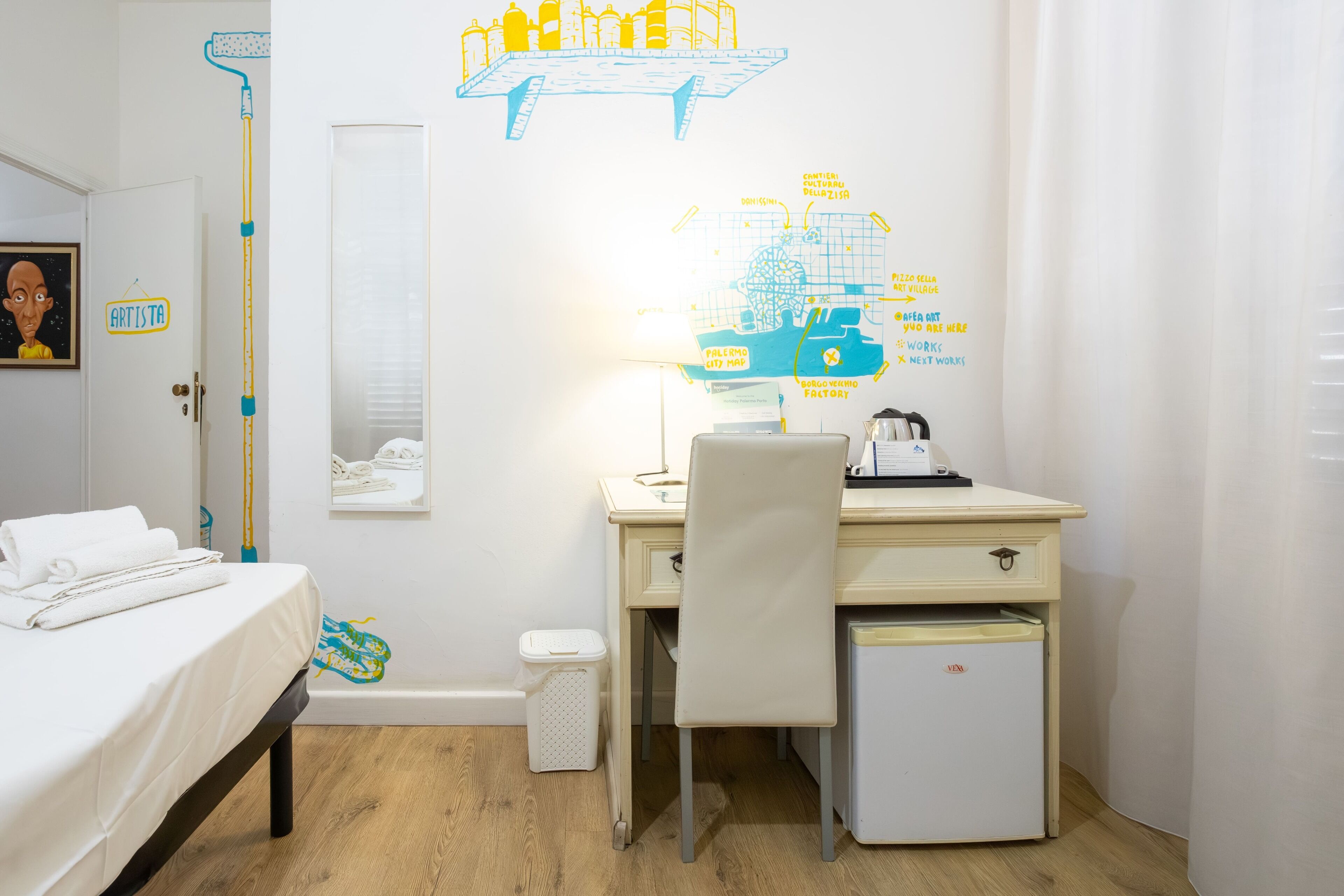 A hotel room with a bed, a desk with a chair, a small refrigerator, and a wall map.