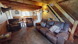 Ein gemütliches Blockhaus-Interieur mit einem Ledersofa, zwei Sesseln, einer Holzbalkendecke und einem aufgehängten Hirschkopf.