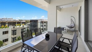 Un balcón con mesa y sillas de vidrio, una silla colgante y vistas a edificios y follaje.