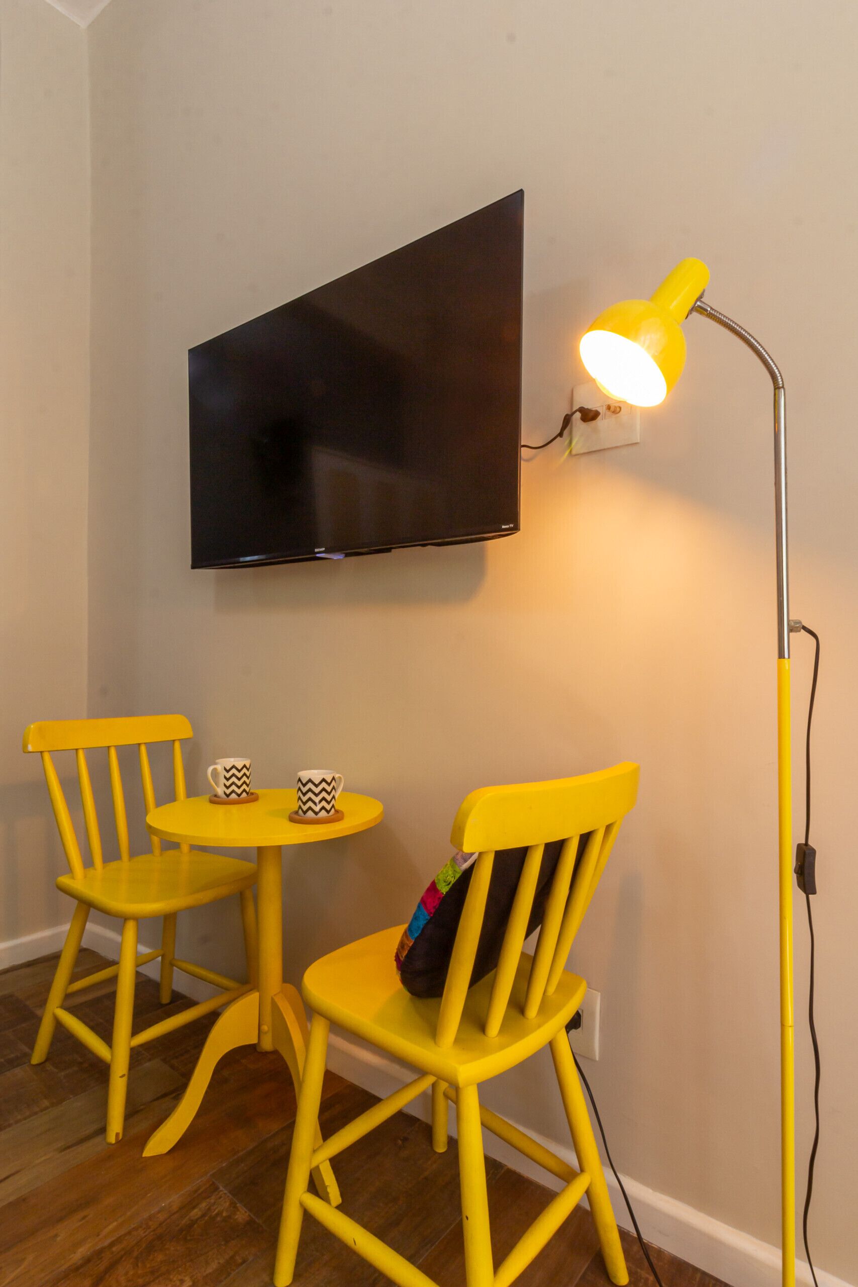 A room with two yellow chairs, a small round table, and a floor lamp. A flat-screen TV is mounted on the wall.