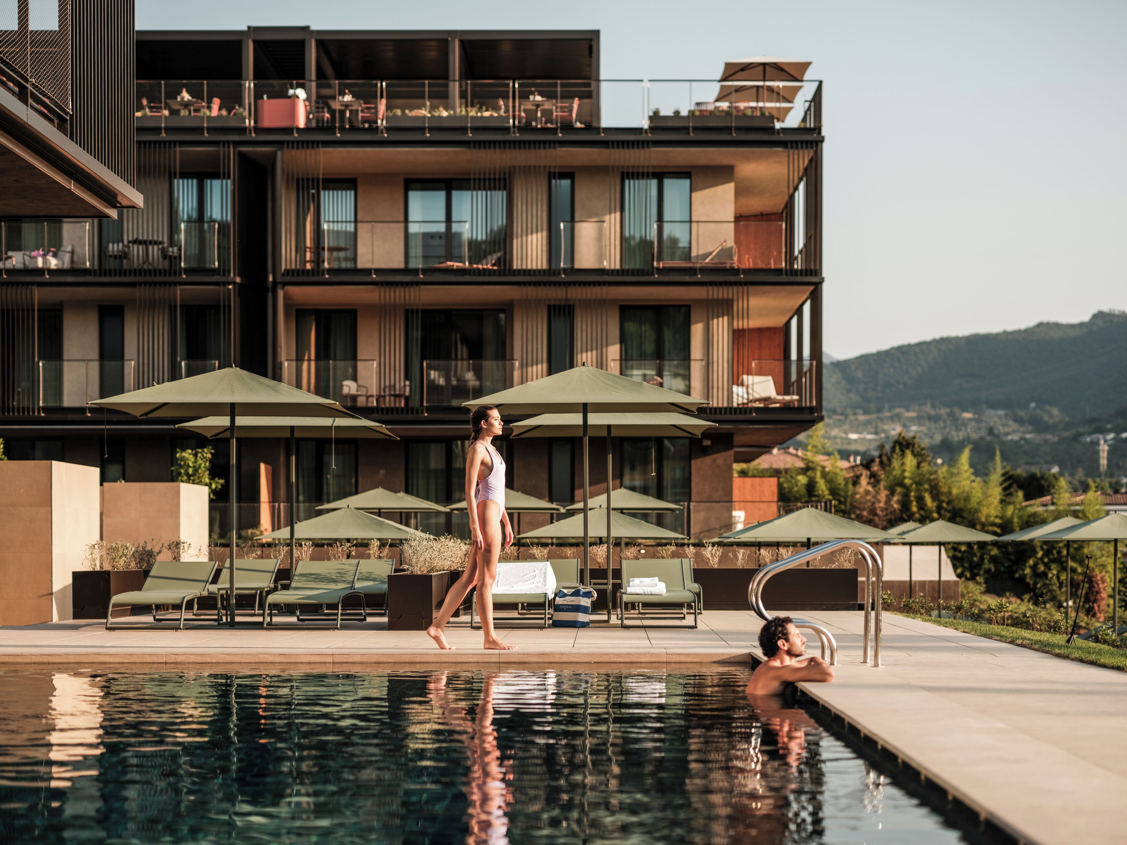 Zona de piscina con sillones de descanso y sombrillas, un edificio moderno con balcones, y vistas del paisaje circundante.