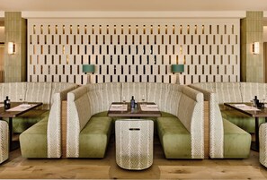 Een restaurant met een modern design, met een uniek wandpatroon, groene bekleding op de stoelen en een centrale eettafel.